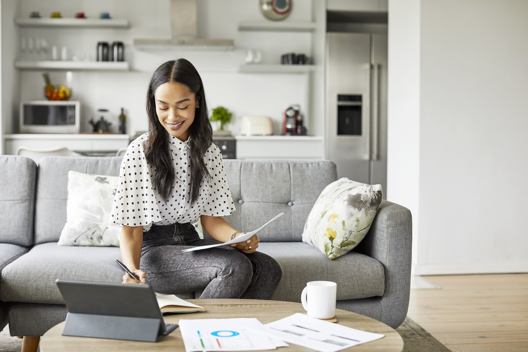 A young woman of color sitting on a couch, looking at financial documents and typing on a tablet.