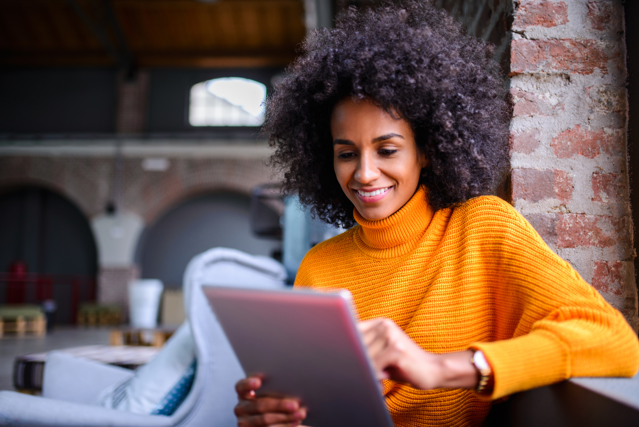 A black woman wearing an orange turtleneck smiles at her rose pink tablet in a coffee shop