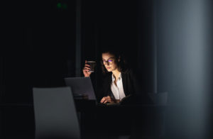 Illustrative image of a white woman in a dark suit sits at a table in a dark office room illuminated by a laptop