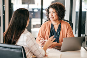 A woman in an orange sweater has a laptop open in front of her while she talks to another woman across a desk