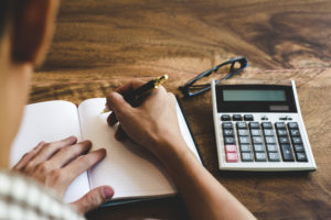 A man holds a pen and writes in a notebook next to a calculator