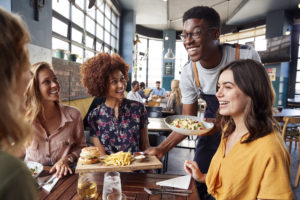 A black male waiter serves a group of black and white women eating at a restaurant