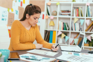 A woman in a yellow sweater sits at a desk covered in financial sheets, typing on a computer