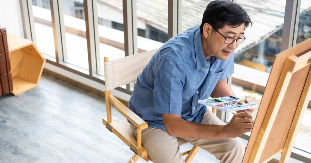 A middle-aged Asian man sits in an artist's chair and paints in a studio