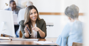 A female banker in an olive green jacket shows an older female customer banking information on her smartphone