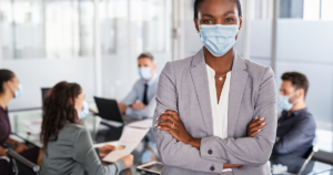 Image of a masked Black businesswoman with her arms crossed standing in front of a table of businessmen. She's looking directly at the camera while they're looking at each other.